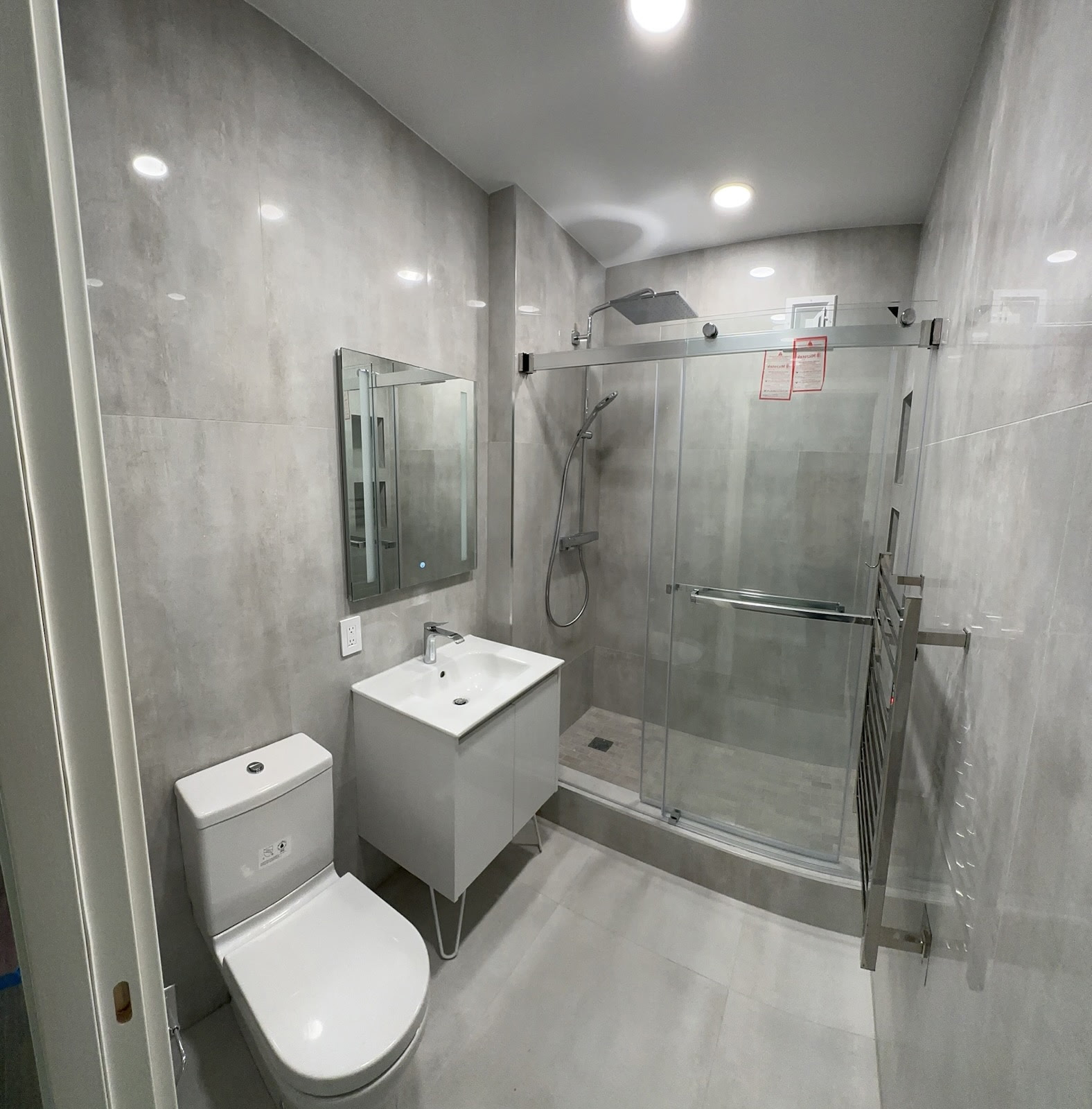 Modern bathroom with grey concrete-look porcelain walls, sliding glass shower enclosure, LED mirror and white vanity on hairpin legs